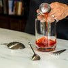 Bell jigger pouring into the A Bar Above mixing glass, with stainless steel julep strainer, spiral bar spoon and cocktail picks.