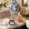 Applying a small amount of Fox Run Craft Wax beneath a blue-and-white porcelain ginger jar, with the open wax container and lid nearby.